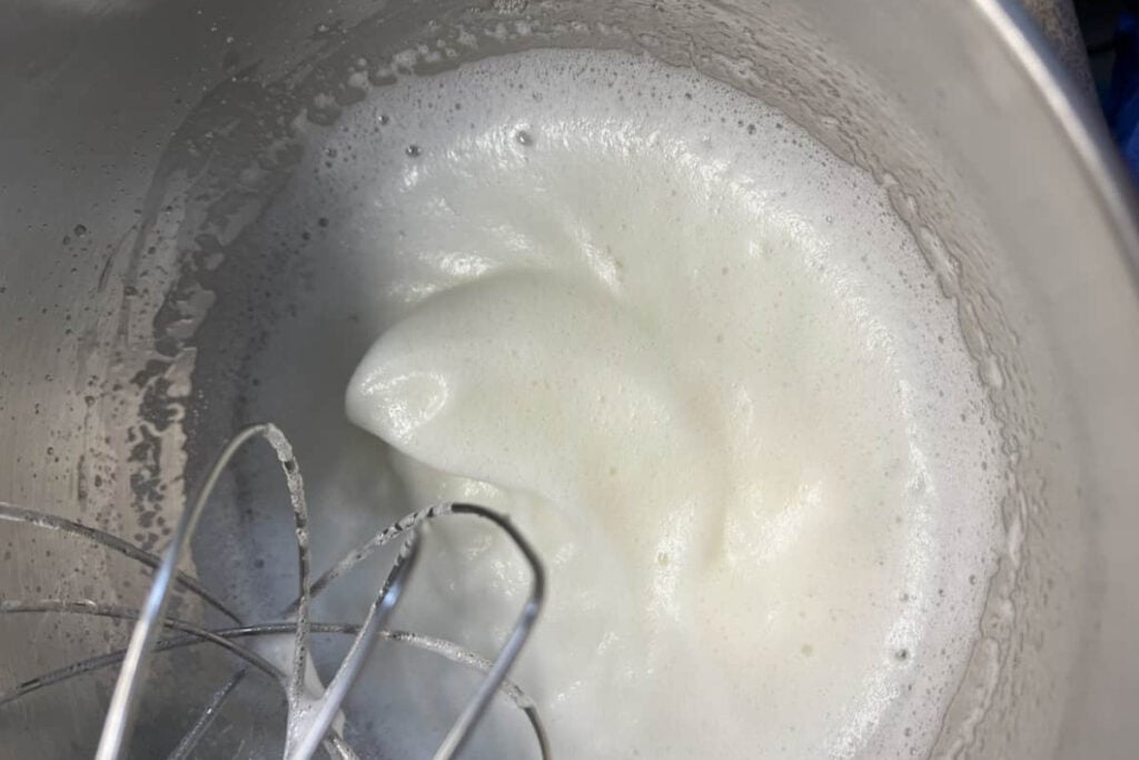 A metal whisk in a bowl with egg whites beaten to soft peaks.