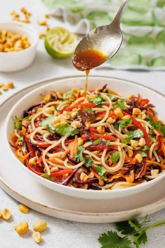 A spoon drizzles sauce over a bowl of colorful noodle salad with peanuts, vegetables, and cilantro on a white plate.
