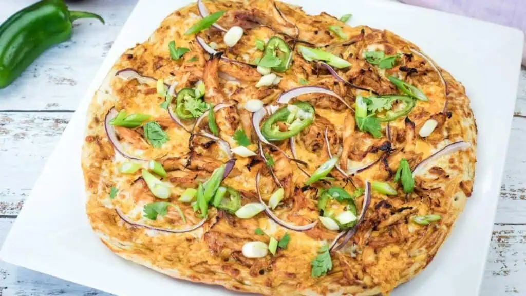 A whole BBQ chicken pizza topped with green onions, cilantro, and jalapeño on a plate.
