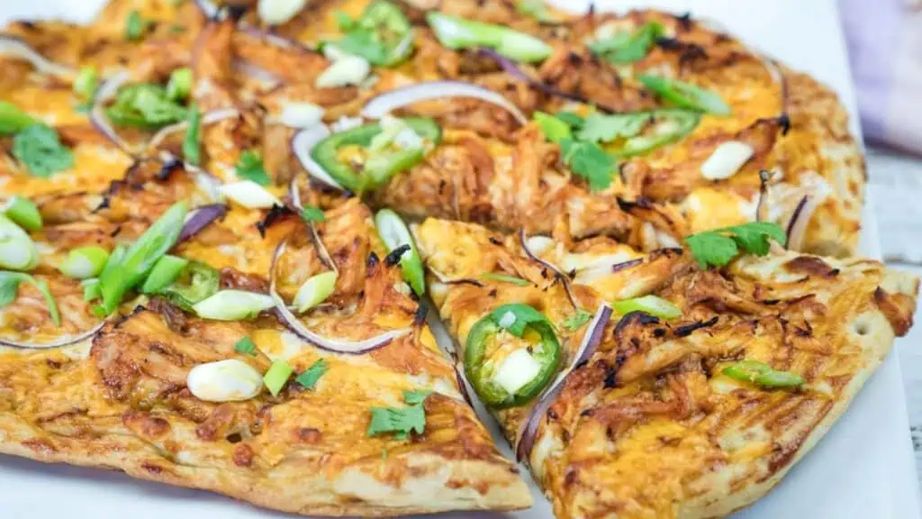 A close-up of a sliced pizza topped with shredded chicken, green onions, red onion slices, jalapeños, cilantro, and melted cheese on a white plate.