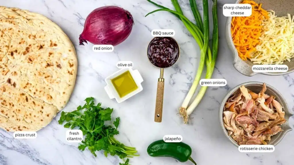 Ingredients for BBQ chicken pizza laid out on a marble surface, including pizza crust, red onion, olive oil, cilantro, BBQ sauce, jalapeño, green onions, cheddar, mozzarella, and chicken.