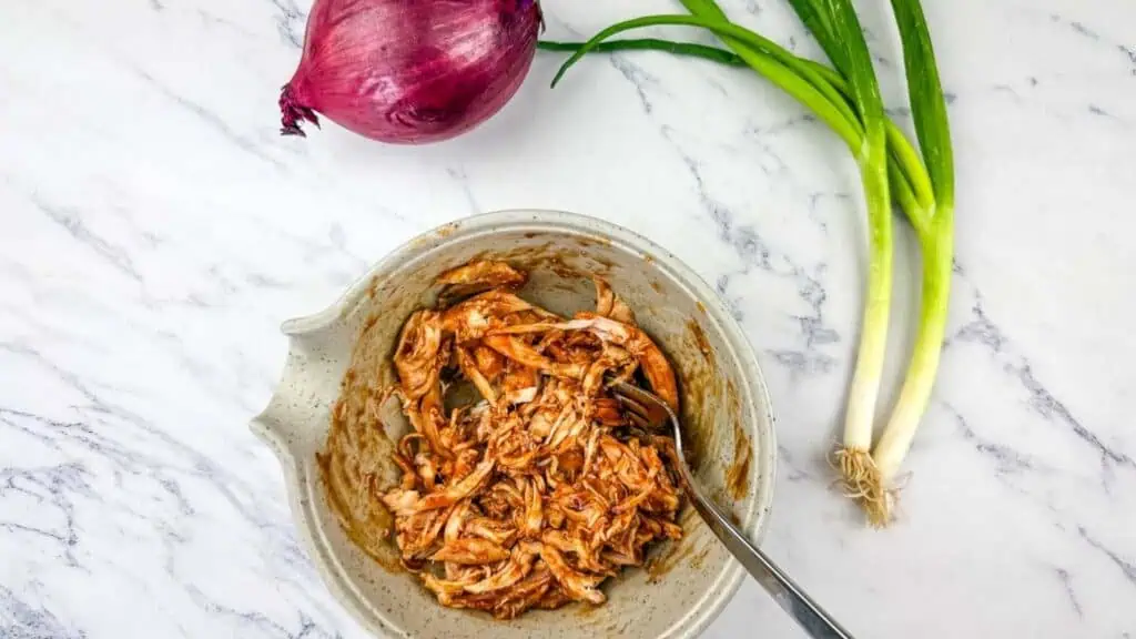 A bowl of shredded chicken mixed with sauce sits on a marble surface next to a red onion and two green onions.