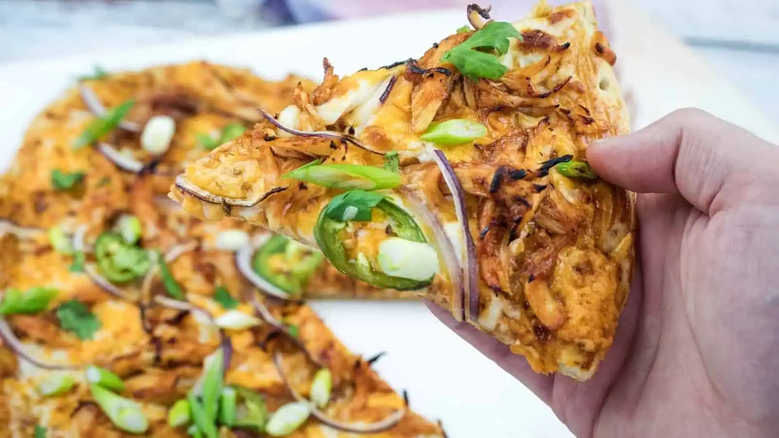 A hand holds a slice of pizza topped with shredded chicken, jalapeño slices, red onions, and green onions; the rest of the pizza is visible on a white surface.