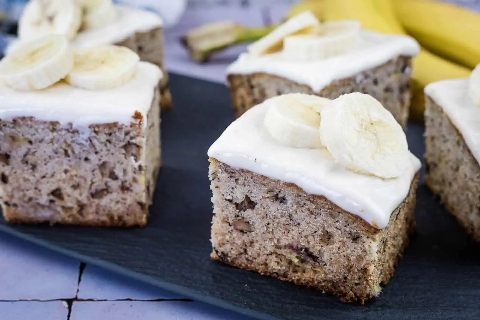Squares of banana cake topped with white frosting and banana slices are arranged on a dark serving tray, with bananas visible in the background.