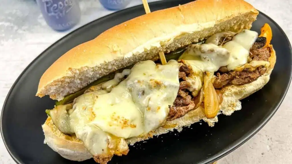 A cheesesteak sandwich with melted cheese, grilled onions, and peppers on a hoagie roll, served on a black plate.