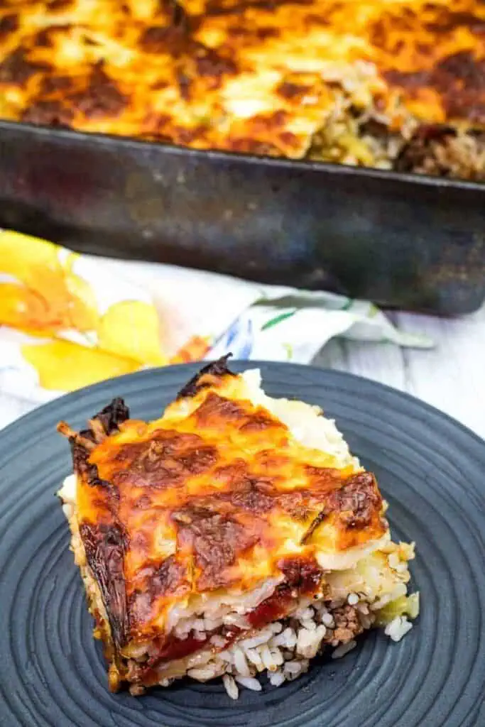 A slice of baked casserole with layers of rice, ground meat, and melted cheese on a plate, with the remaining casserole in a pan in the background.