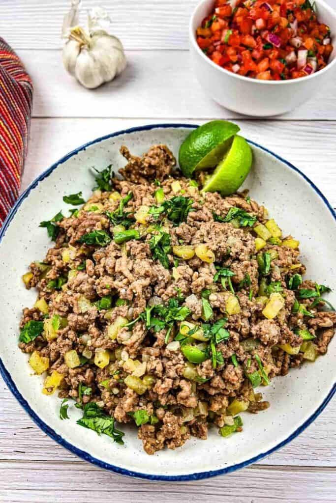 A bowl of cooked ground beef mixed with diced green peppers and herbs, garnished with cilantro and lime wedges, with a side of salsa and garlic in the background.