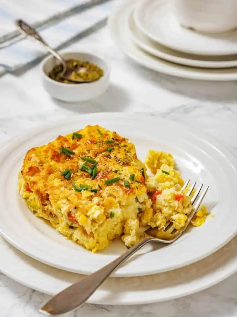A serving of baked corn pudding casserole with herbs on a white plate, accompanied by a fork; extra sauce in a small bowl and stacked plates are in the background.