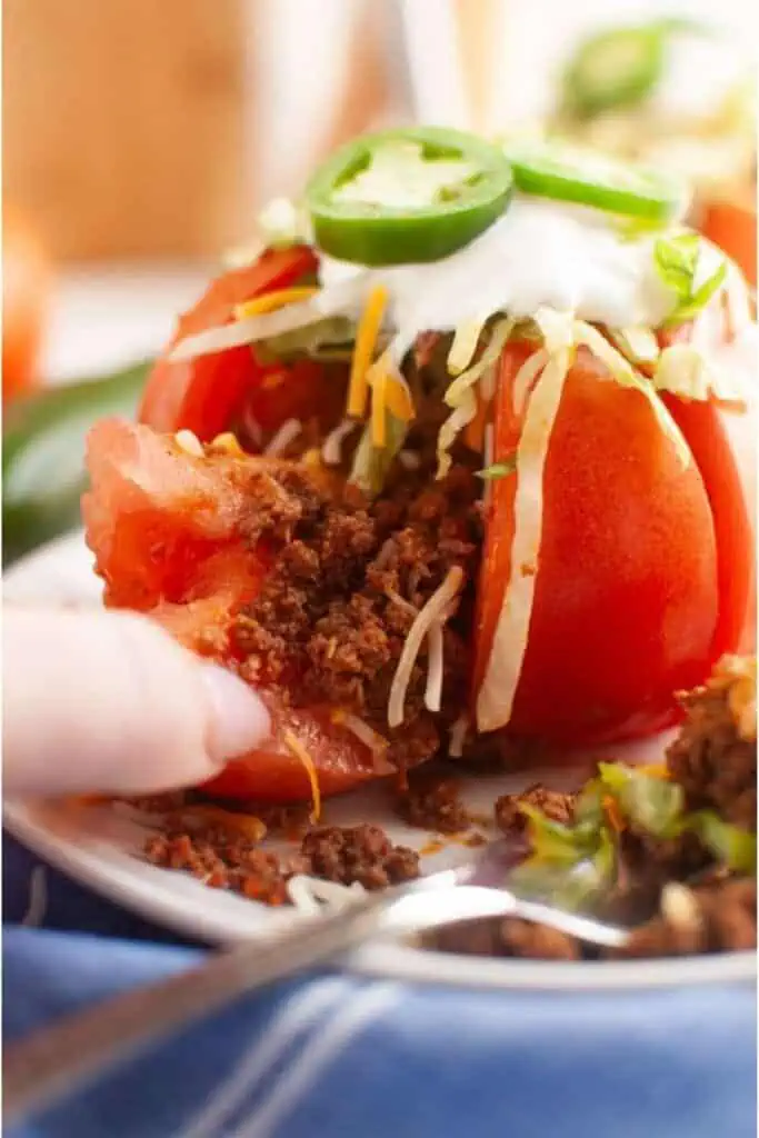 A halved tomato stuffed with ground beef, shredded cheese, lettuce, and topped with sour cream and sliced jalapeños on a plate.