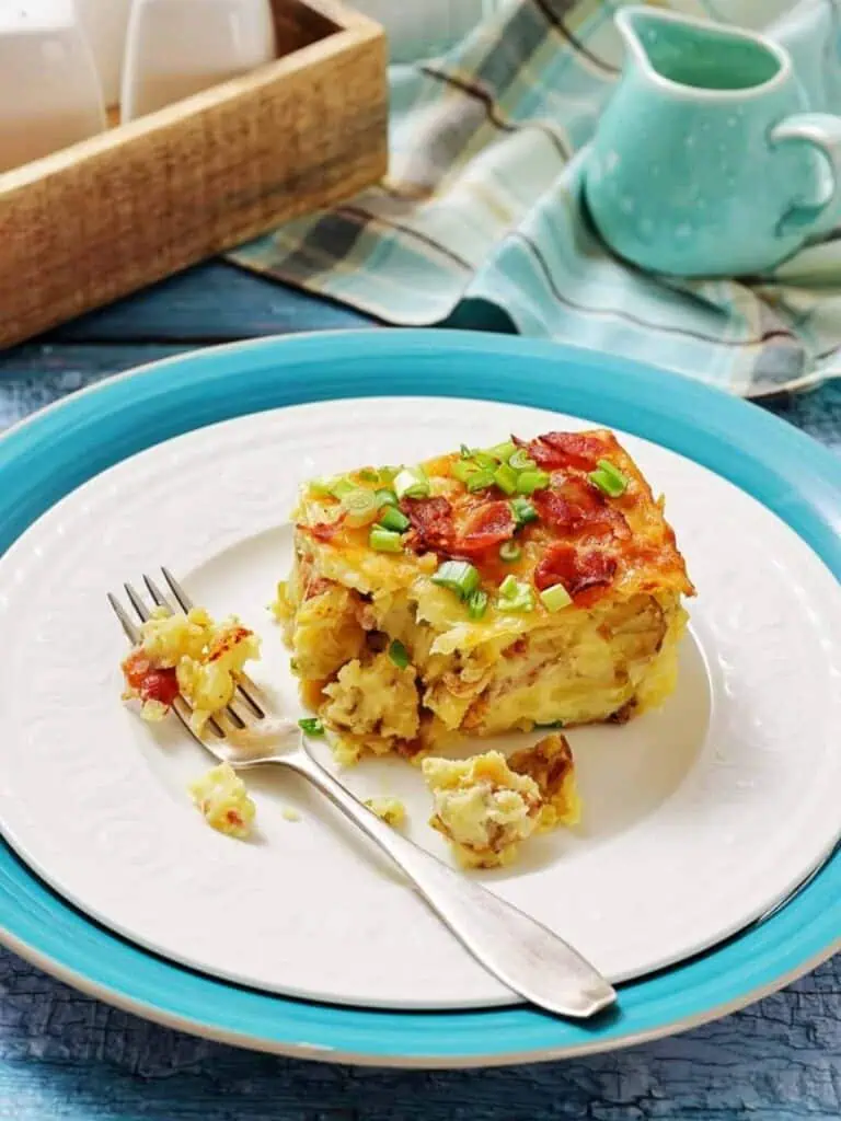 A slice of breakfast casserole topped with chopped green onions and bacon sits on a white plate with a fork, on a blue charger, next to a blue pitcher and a wooden tray.