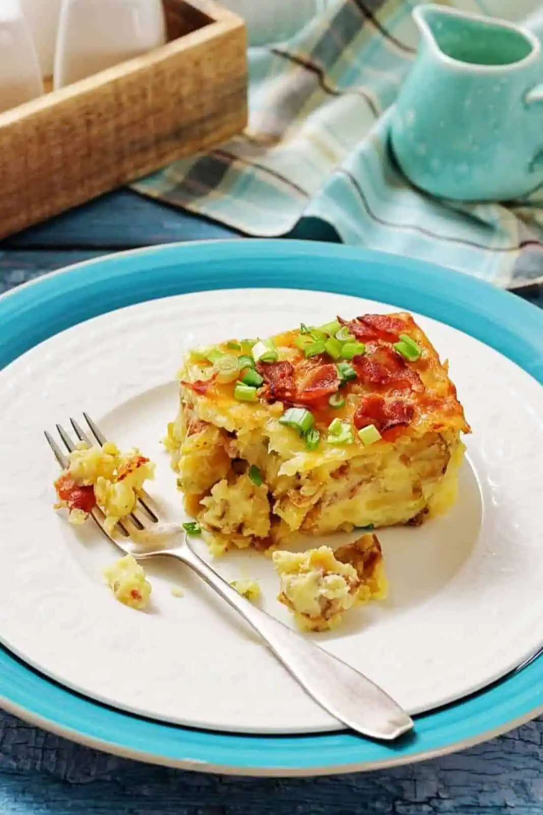 A slice of breakfast casserole topped with chopped green onions and bacon sits on a white plate with a fork, on a blue charger, next to a blue pitcher and a wooden tray.