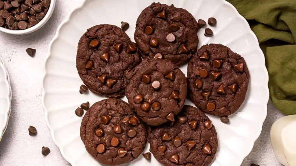 A white plate displays six chocolate cookies with chocolate chips, surrounded by a green cloth and a bowl of chocolate chips.