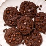 Six Copycat Crumbl Dark Dream Cookies are arranged on a white scalloped plate, with extra chocolate chips scattered around.