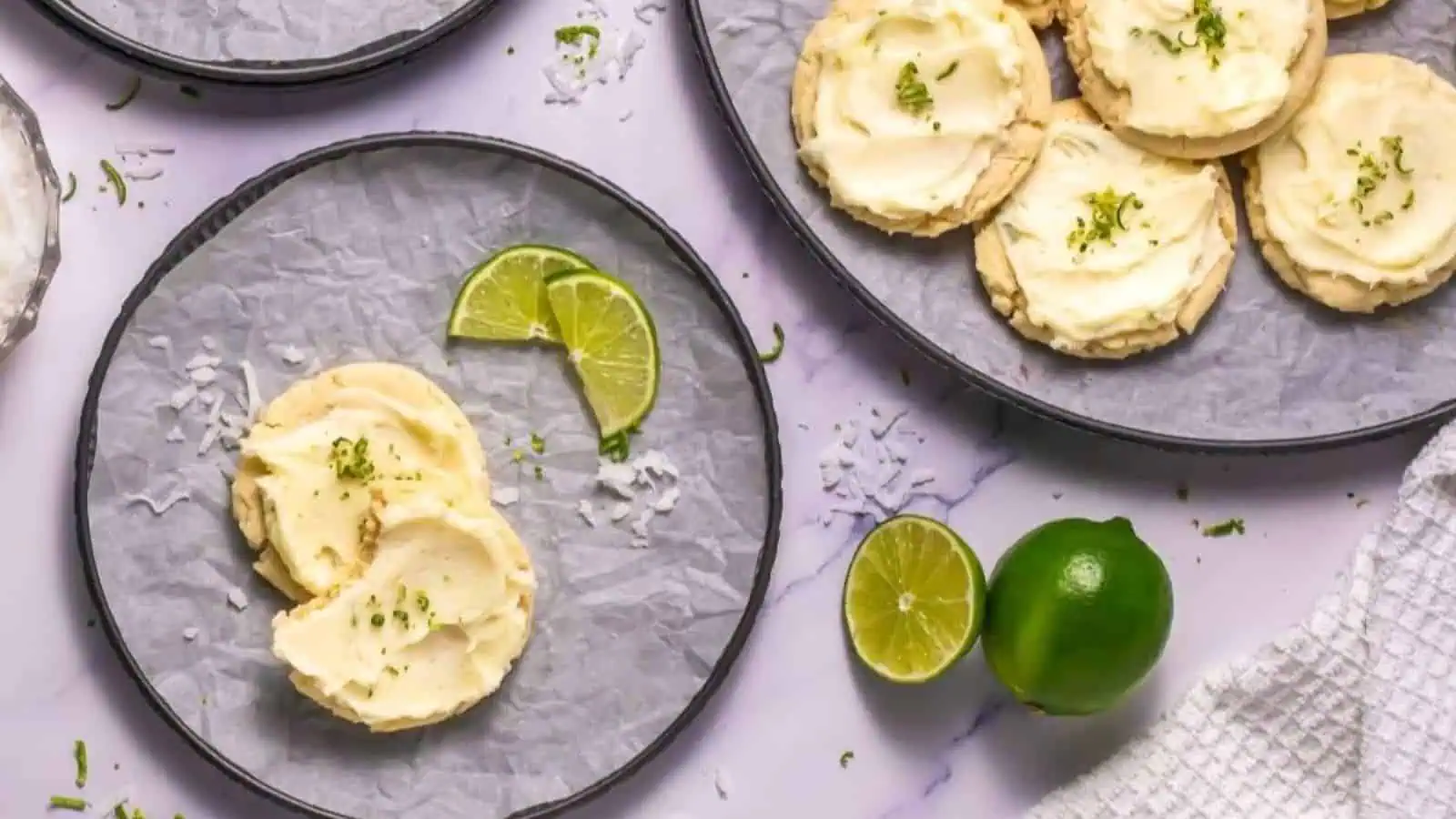 Plates of frosted cookies garnished with lime zest, with lime slices and a whole lime on a marble surface.