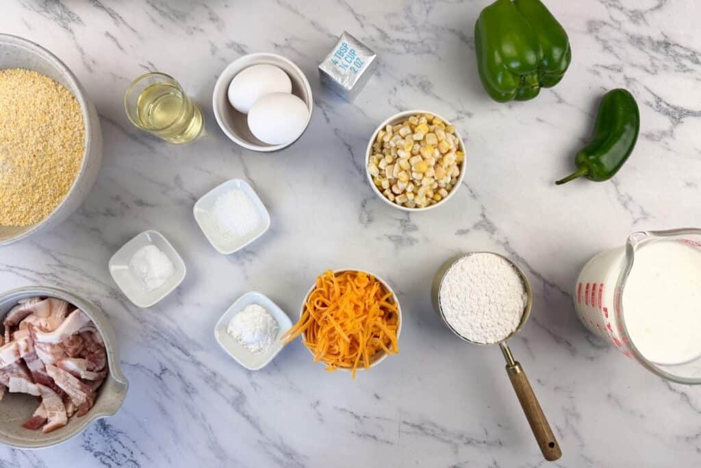 Ingredients for a recipe arranged on a marble countertop, including eggs, cornmeal, flour, shredded cheese, corn, bacon, milk, green peppers, oil, butter, and small bowls of powders.