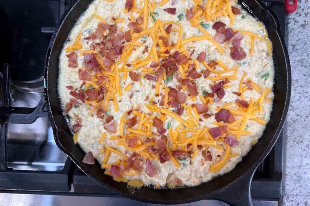 A cast iron skillet on a stove filled with a creamy mixture topped with shredded cheddar cheese and crumbled bacon.