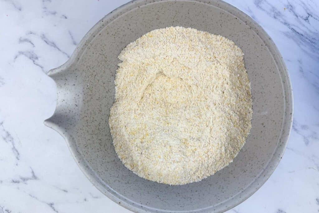 A speckled grey mixing bowl filled with a mound of dry cornmeal sits on a white marble surface.