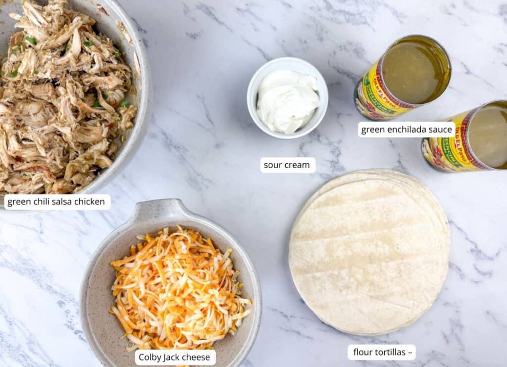 Flat lay of labeled enchilada ingredients: green chili salsa chicken, Colby Jack cheese, sour cream, green enchilada sauce, and flour tortillas on a marble surface.