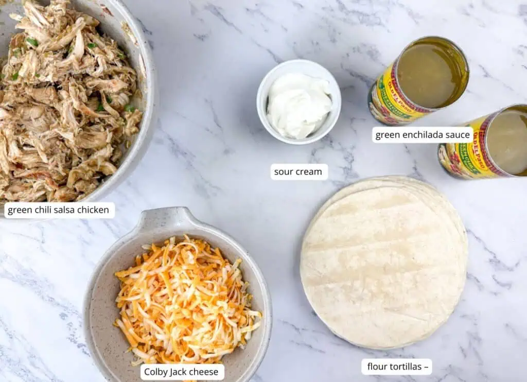 Flat lay of labeled enchilada ingredients: green chili salsa chicken, Colby Jack cheese, sour cream, green enchilada sauce, and flour tortillas on a marble surface.