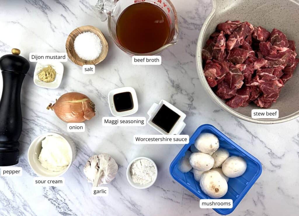 Ingredients for beef stroganoff, including stew beef, beef broth, mushrooms, onion, garlic, sour cream, Dijon mustard, Worcestershire sauce, Maggi seasoning, salt, pepper, and flour.