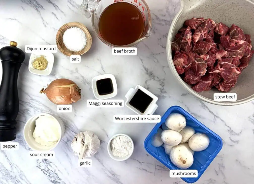 Ingredients for beef stroganoff, including stew beef, beef broth, mushrooms, onion, garlic, sour cream, Dijon mustard, Worcestershire sauce, Maggi seasoning, salt, pepper, and flour.