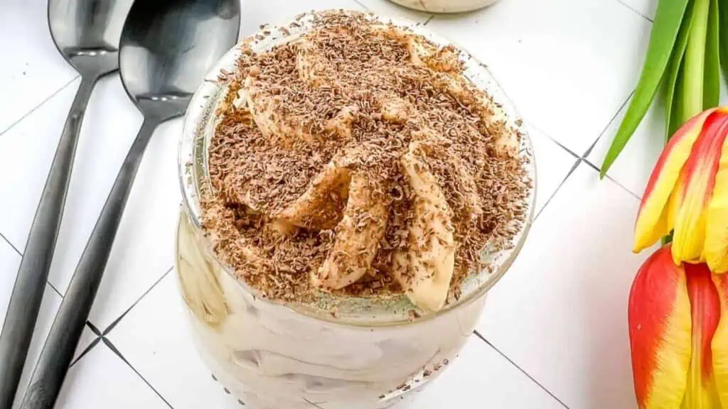A glass of creamy tiramisu topped with chocolate shavings is placed on a white tiled surface next to two spoons and a red-yellow tulip.