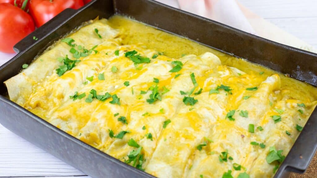 A baking dish filled with enchiladas topped with melted cheese and chopped cilantro, with tomatoes in the background.