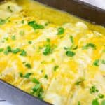 A baking dish filled with cheesy enchiladas topped with melted cheese and chopped cilantro on a white wooden surface.