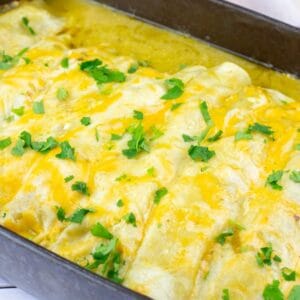 A baking dish filled with cheesy enchiladas topped with melted cheese and chopped cilantro on a white wooden surface.
