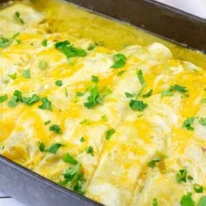 A baking dish filled with cheesy enchiladas topped with melted cheese and chopped cilantro on a white wooden surface.