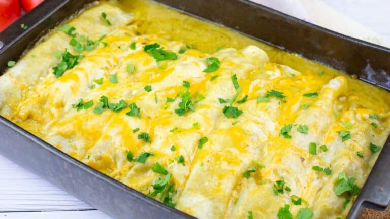 A baking dish filled with cheesy enchiladas topped with melted cheese and chopped cilantro on a white wooden surface.