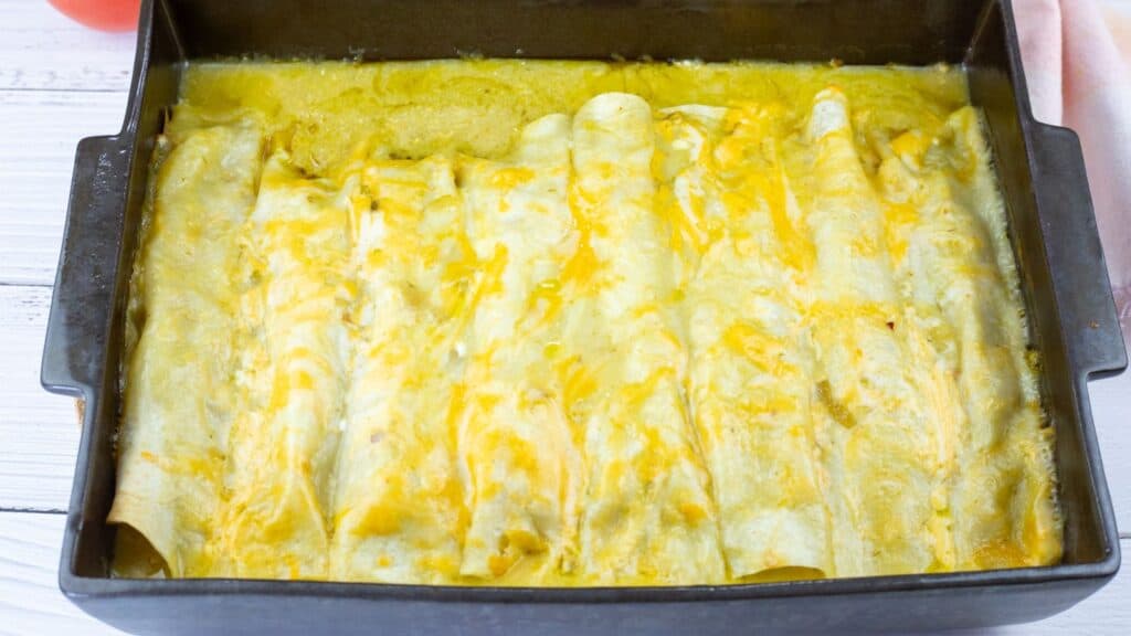 A baking dish filled with enchiladas covered in melted cheese, fresh out of the oven, on a white wooden surface.