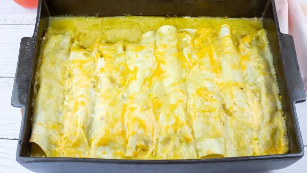 A baking dish filled with enchiladas covered in melted cheese, fresh out of the oven, on a white wooden surface.