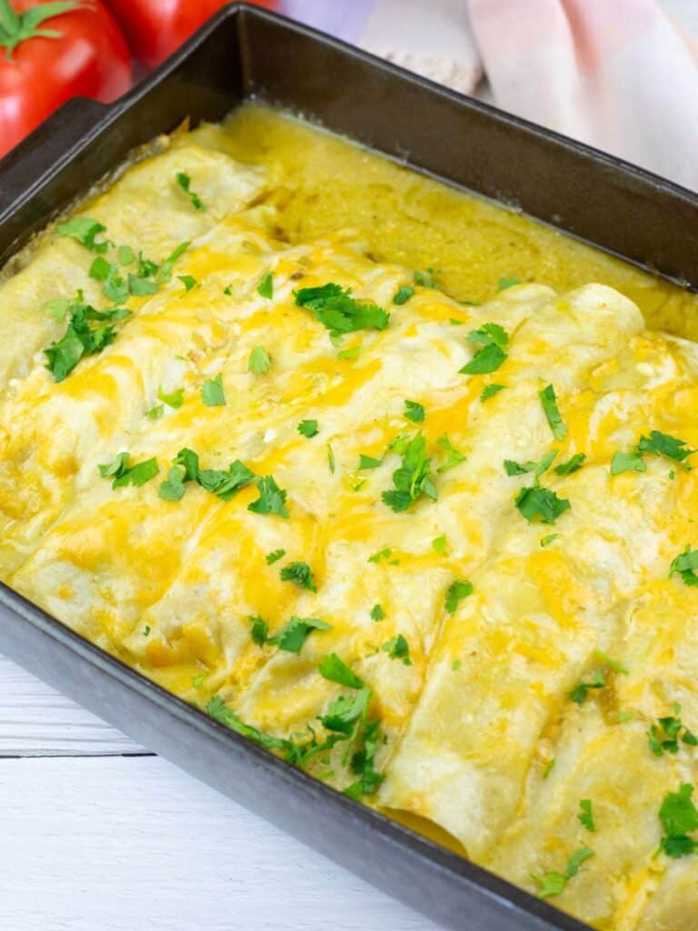 A baking dish filled with enchiladas topped with melted cheese and chopped cilantro, placed on a white wooden surface.