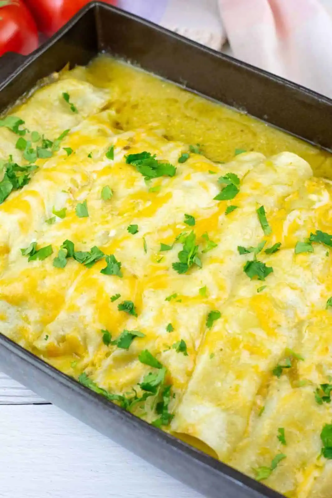 A baking dish filled with enchiladas topped with melted cheese and chopped cilantro, placed on a white wooden surface.
