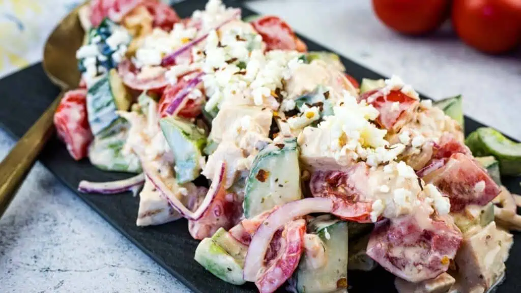 A plate of chopped salad with cucumbers, tomatoes, red onions, feta cheese, and creamy dressing on a dark rectangular serving dish.