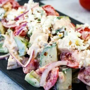 A plate of chopped salad with cucumbers, tomatoes, red onions, feta cheese, and creamy dressing on a dark rectangular serving dish.