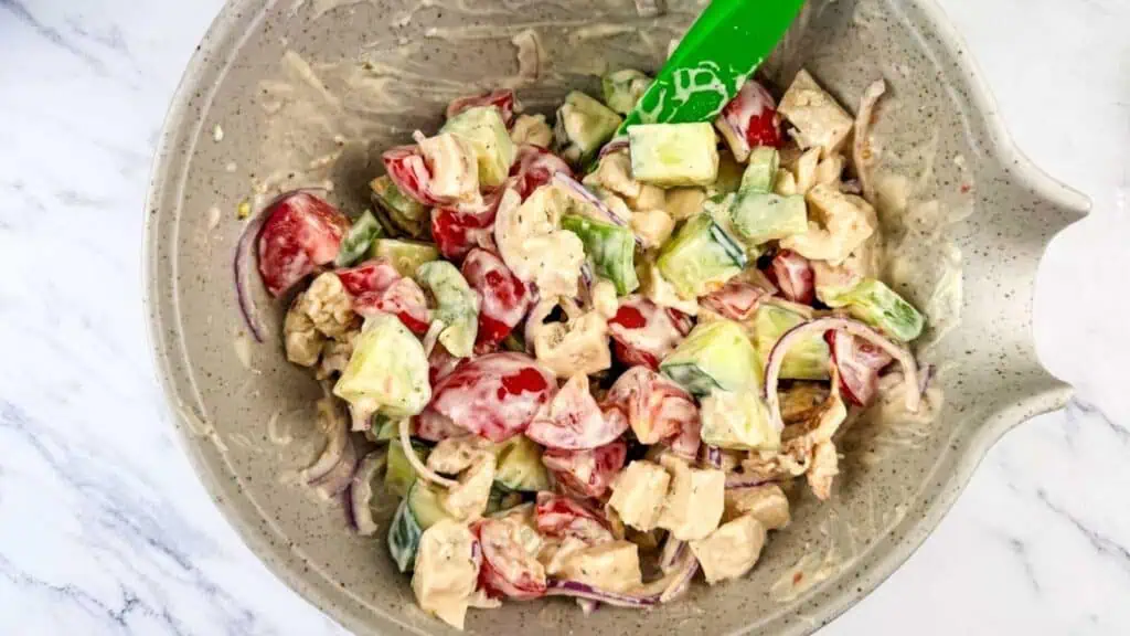A bowl of mixed salad with chopped cucumbers, tomatoes, red onions, and pieces of chicken coated in a creamy dressing, with a green spatula.