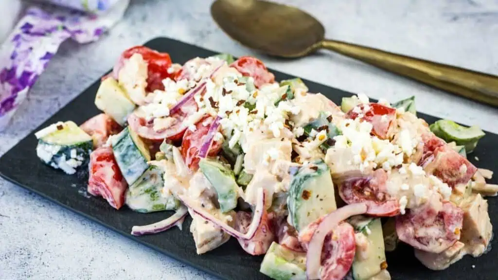 A plate of Greek salad with chopped tomatoes, cucumbers, red onion, feta cheese, and creamy dressing on a dark serving board, with a spoon in the background.