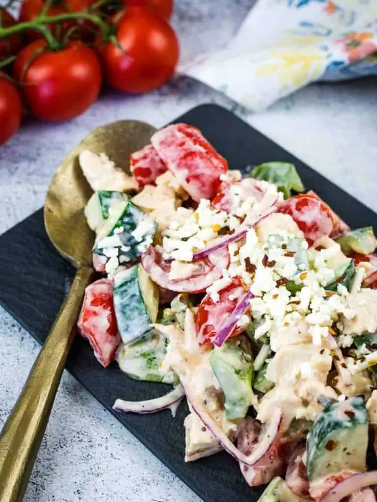 A plate of chopped salad with tomatoes, cucumbers, red onions, chicken, crumbled feta cheese, and creamy dressing, served with a gold spoon.