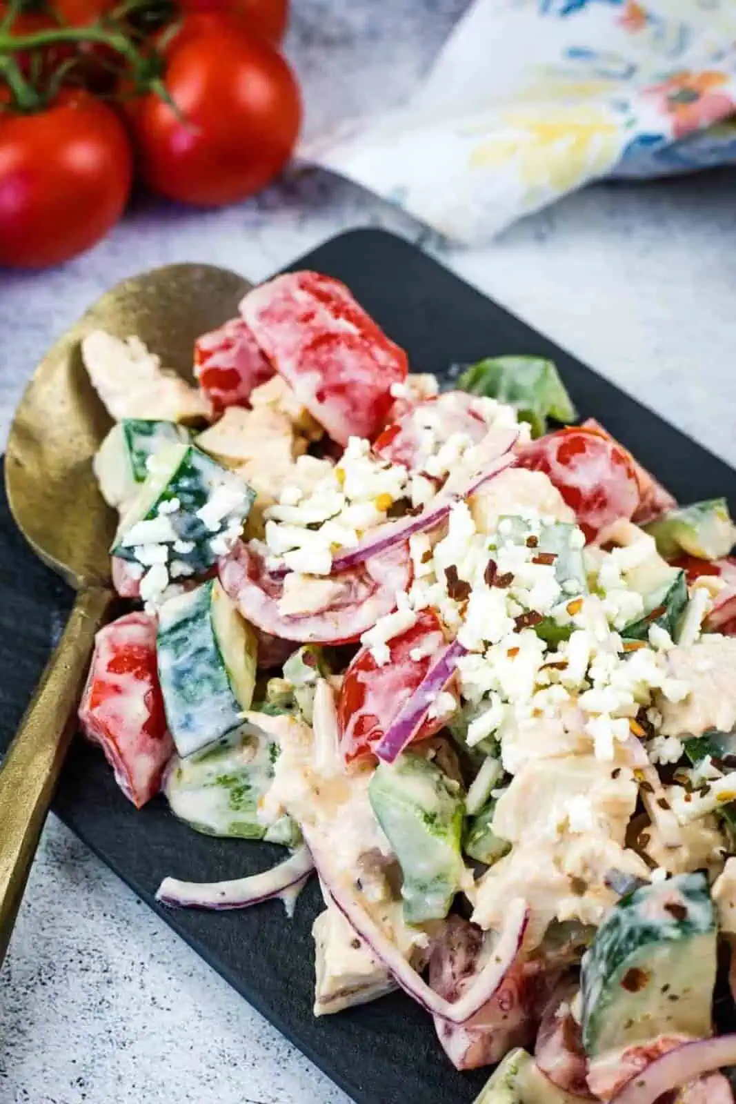 A plate of chopped salad with tomatoes, cucumbers, red onions, chicken, crumbled feta cheese, and creamy dressing, served with a gold spoon.