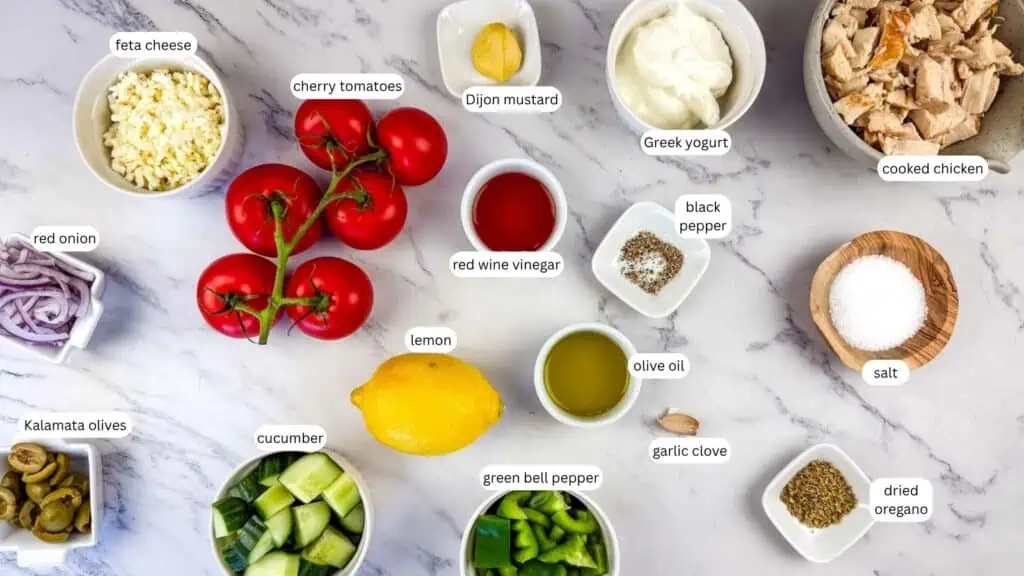 Ingredients for a Greek salad are arranged on a marble surface, each labeled, including feta cheese, cherry tomatoes, red onion, olives, cucumber, and cooked chicken.