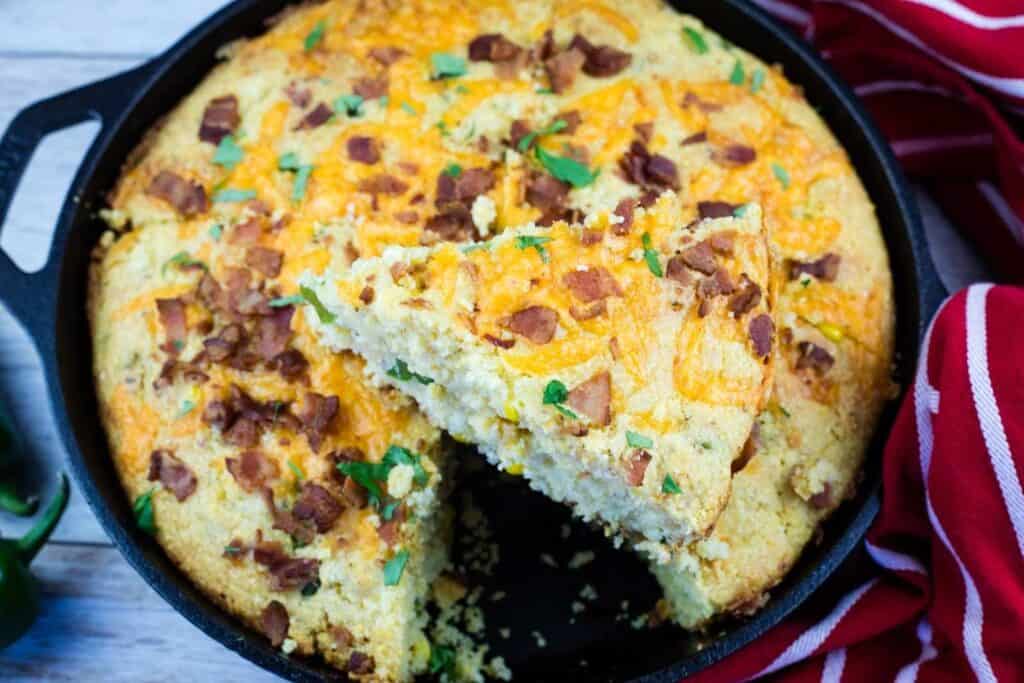 A skillet of baked cornbread topped with cheese, bacon pieces, and herbs, with one triangular slice being lifted out.