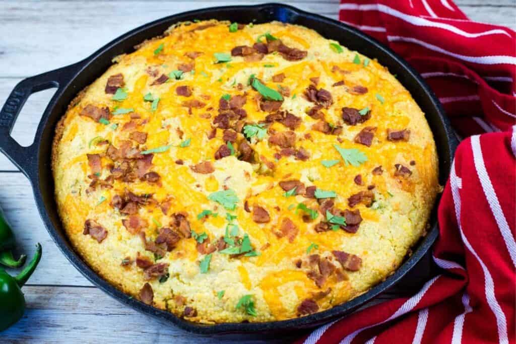 A cast-iron skillet filled with baked cornbread topped with chopped bacon and herbs, placed next to a red striped kitchen towel.