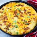 A cast-iron skillet filled with baked cornbread topped with chopped bacon and herbs, placed next to a red striped kitchen towel.