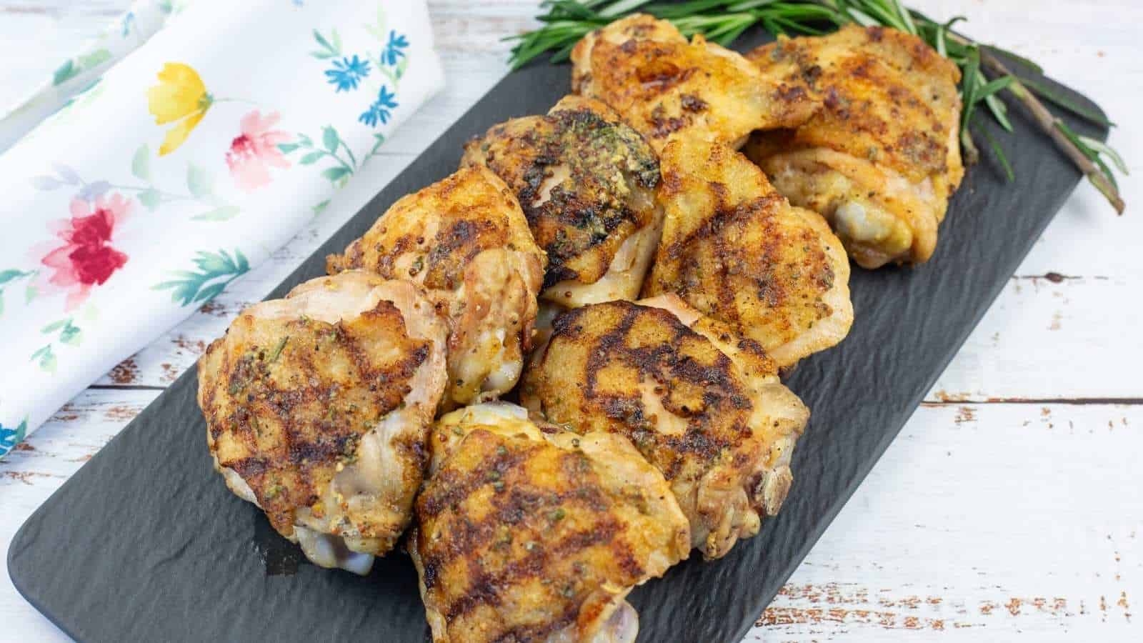 Grilled Dijon rosemary chicken thighs are arranged on a rectangular black slate platter, garnished with fresh rosemary, next to a white floral napkin on a wooden surface.