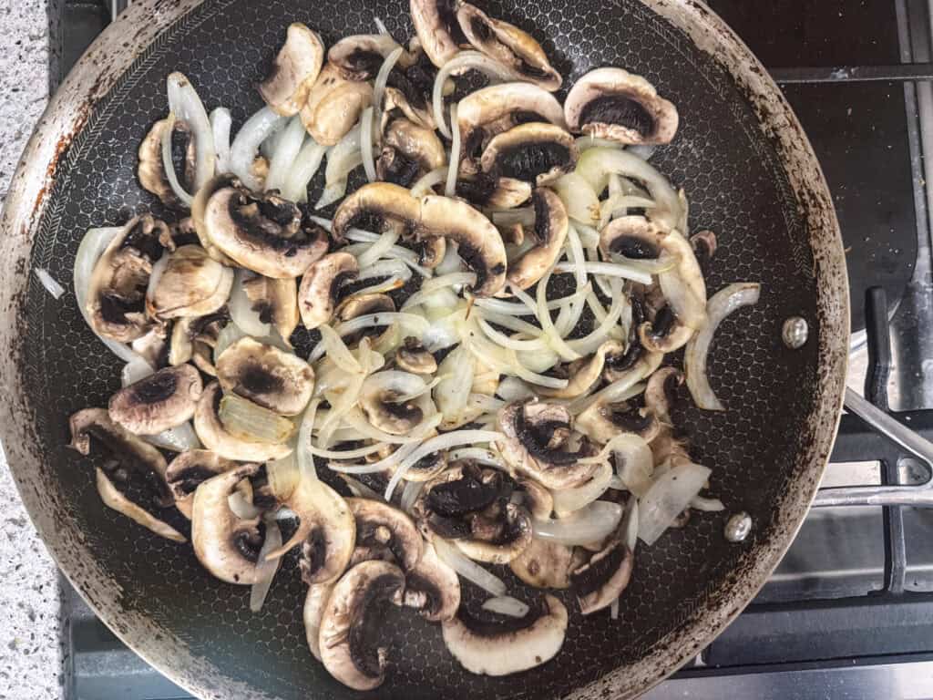 Sliced mushrooms and onions are being saut&eacute;ed in a frying pan on a stovetop.