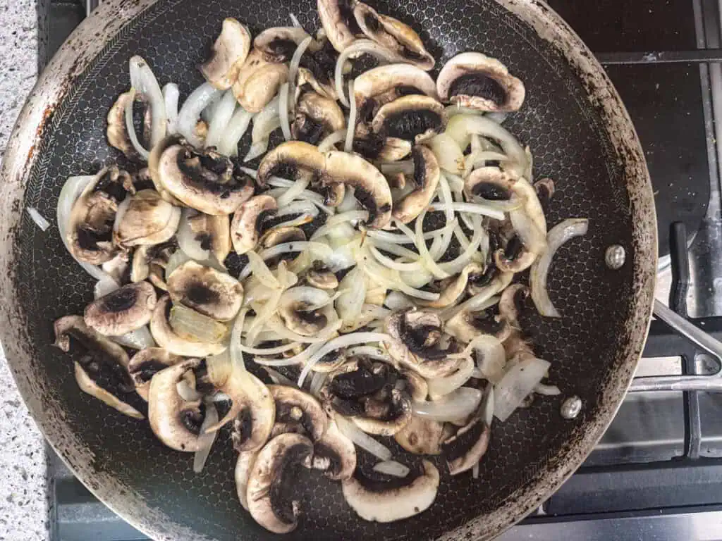 Sliced mushrooms and onions are being sautéed in a frying pan on a stovetop.