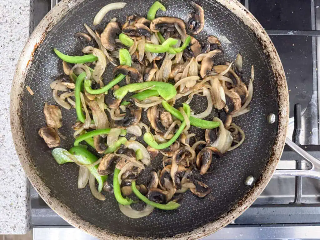 Sliced mushrooms, green bell peppers, and onions cooking in a frying pan on a stovetop.