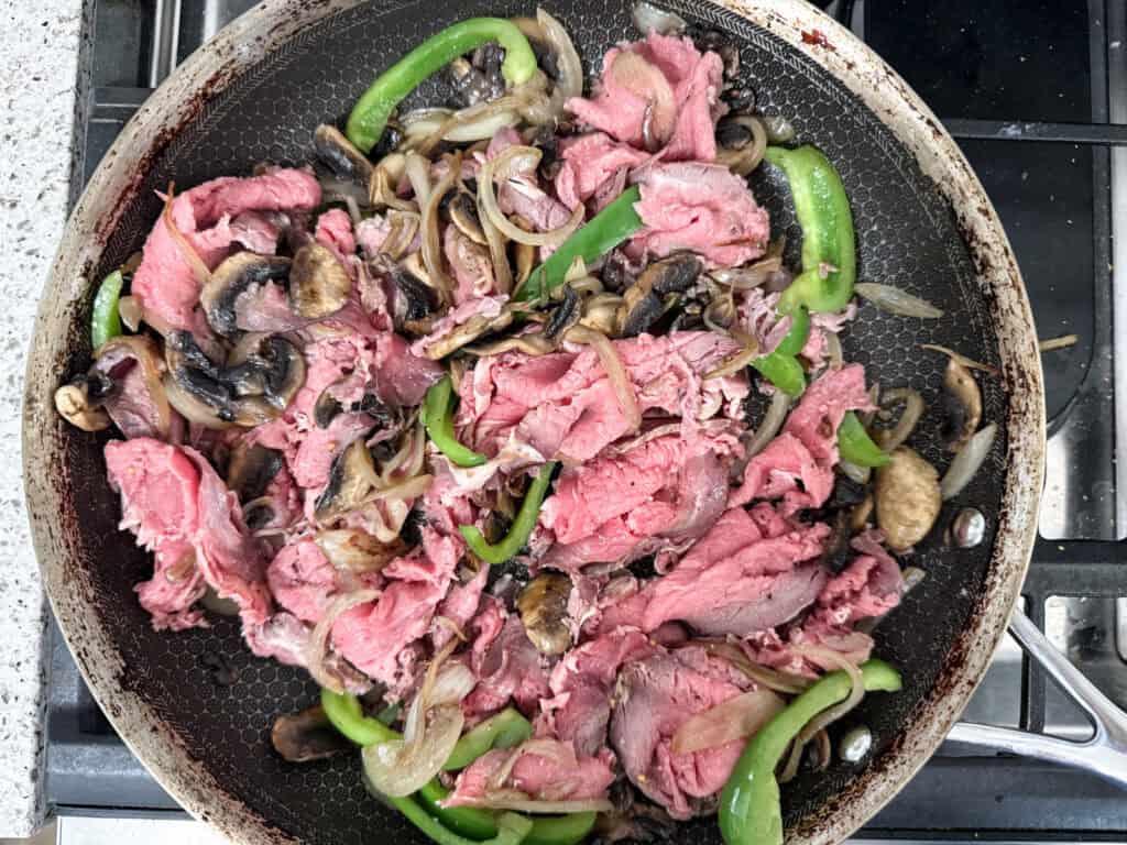 Sliced raw beef, green bell peppers, onions, and mushrooms cooking in a frying pan on a stovetop.